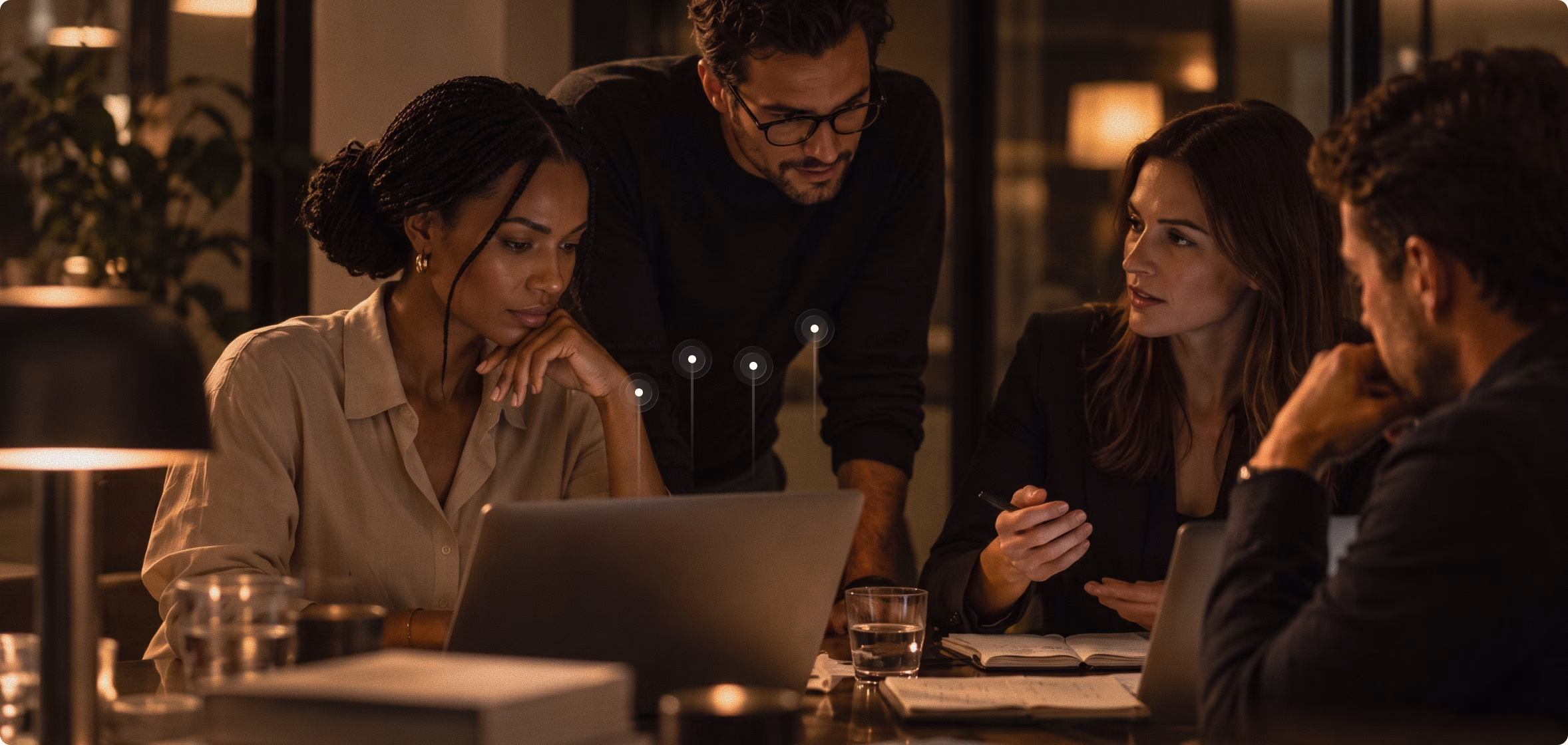 Four colleagues around a table in a dim office at night: one studies a laptop with her chin on her hand while the others lean in and talk it through.